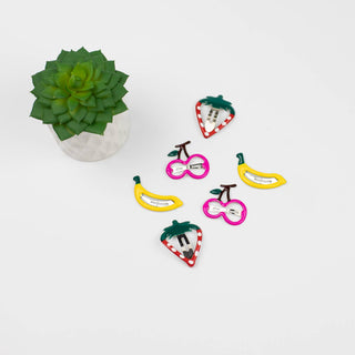 Colorful fruit-shaped hair clips on a white surface with a small potted plant.