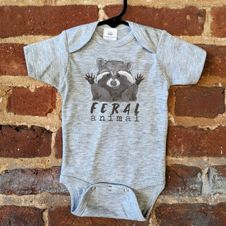 Gray baby onesie with raccoon graphic and 'Feral animal' text on a brick wall background