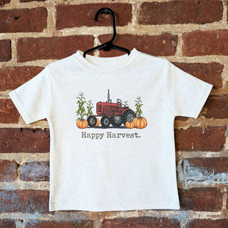 White t-shirt with a red tractor and pumpkins design, labeled 'Happy Harvest', on a hanger against a brick wall.