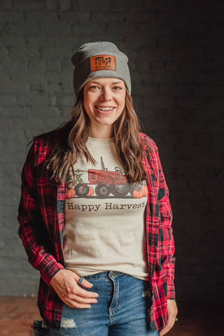 Person wearing a red plaid shirt, white t-shirt with a graphic design, and gray cap against a dark brick wall.