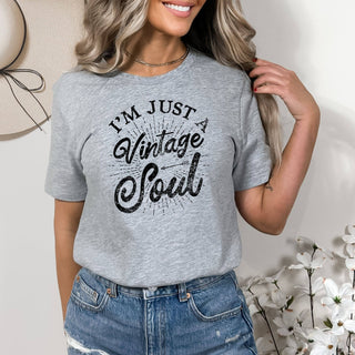 Woman wearing a gray t-shirt with 'I'm Just a Vintage Soul' text, standing against a neutral background with flowers.