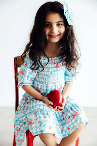 Young girl in a patterned dress holding a red heart-shaped object, sitting on a chair against a white background.