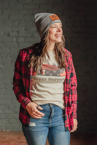 Person wearing a plaid shirt, beanie, and 'Happy Harvest' t-shirt against a brick wall.