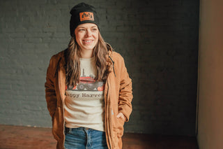 Person wearing a brown jacket, white t-shirt with graphics, and black beanie with text against a dark brick wall.